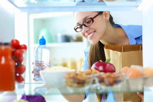 Portret van een vrouw in de buurt van een open koelkast vol gezond voedsel, groenten en fruit. — Stockfoto