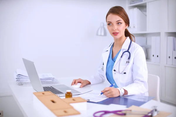 Hermosa joven sonriente doctora sentada en el escritorio y escribiendo —  Fotos de Stock