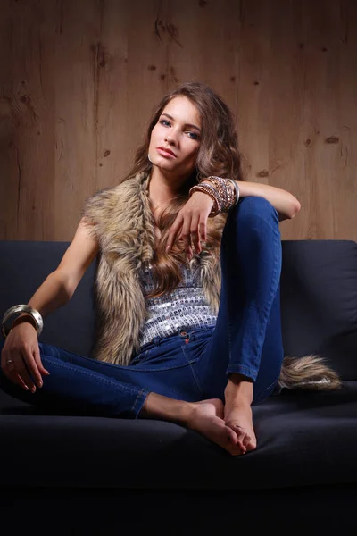 Retrato de mulher elegante sentado no sofá preto vestindo uma calça azul e colete de pele — Fotografia de Stock