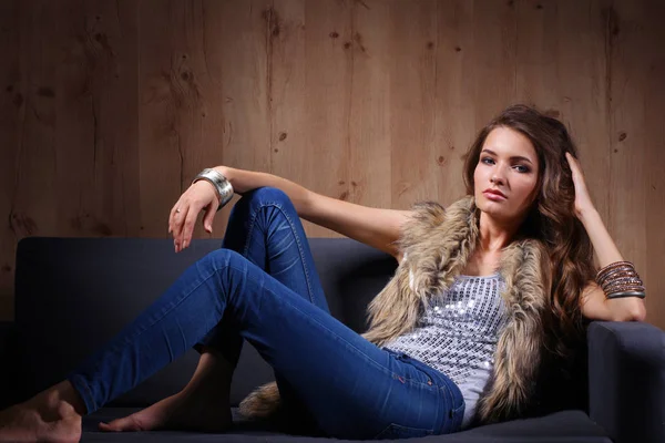 Portrait of elegant woman sitting on black sofa wearing a blue jeans and fur vest Stock Picture