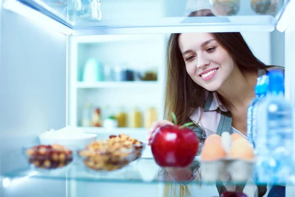 Portret van een vrouw in de buurt van een open koelkast vol gezond voedsel, groenten en fruit. — Stockfoto