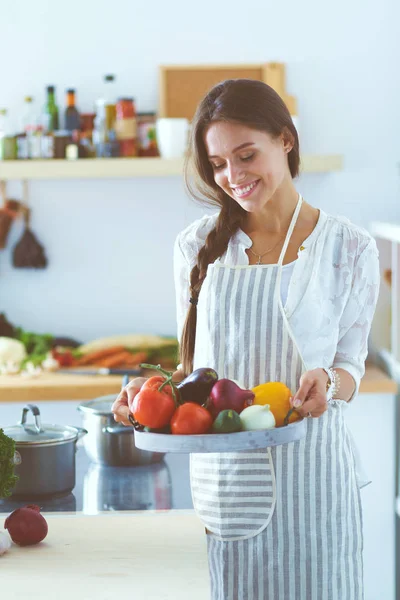 Mujer joven sonriente sosteniendo verduras de pie en la cocina. Jovencita sonriente —  Fotos de Stock