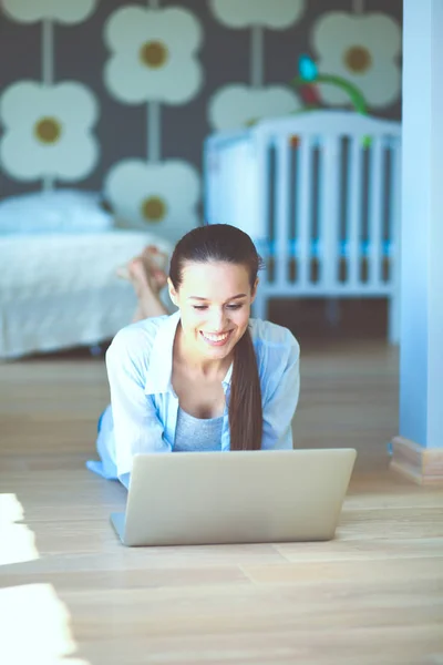 Ung kvinna som sitter på golvet nära barnens babysäng med laptop. Ung mamma — Stockfoto