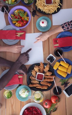 Ahşap masada otururken birlikte yemek yiyen bir grup insanın üst düzey görüntüsü. Masada yemek var. İnsanlar fast food yiyor..