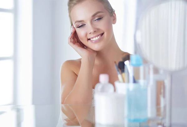 Mooie jonge vrouw is aanraken haar gezicht en glimlachen terwijl kijken naar de spiegel — Stockfoto