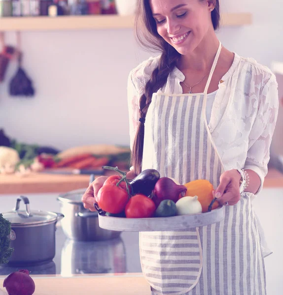 Mujer joven sonriente sosteniendo verduras de pie en la cocina. Jovencita sonriente —  Fotos de Stock