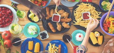 Ahşap masada otururken birlikte yemek yiyen bir grup insanın üst düzey görüntüsü. Masada yemek var. İnsanlar fast food yiyor..