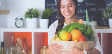 Sebzeli alışveriş poşeti taşıyan genç bir kadın. Mutfakta duruyor.