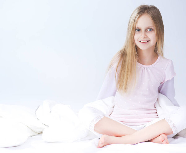 Little girl in the bedroom is sitting on the bed. Little girl is wearing a pajamas and sitting in bed