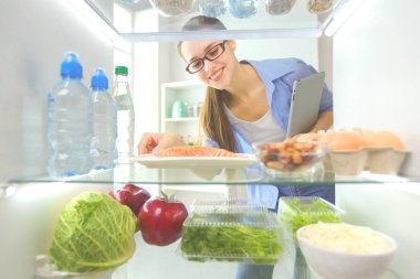 Sağlıklı yiyecek, sebze ve meyvelerle dolu açık buzdolabının yanında duran kadın portresi..