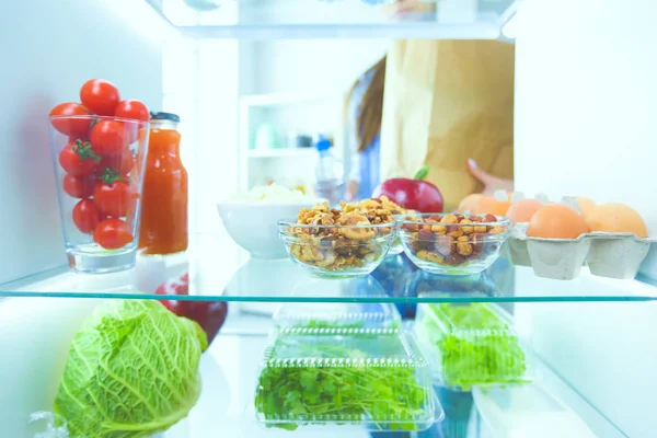 Портрет женщины, стоящей рядом с открытым холодильником, полным здоровой пищи, овощей и фруктов. — стоковое фото