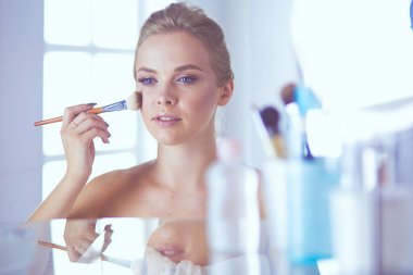 Güzel genç bir kadın yüzüne dokunuyor ve aynaya bakarken gülümsüyor.