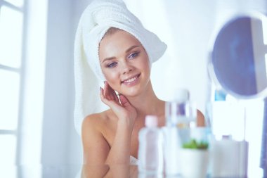 Güzel genç bir kadın yüzüne dokunuyor ve aynaya bakarken gülümsüyor.