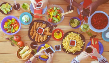 Ahşap masada otururken birlikte yemek yiyen bir grup insanın üst düzey görüntüsü. Masada yemek var. İnsanlar fast food yiyor..