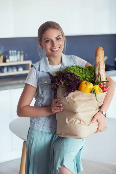 Sebzeli alışveriş poşeti taşıyan genç bir kadın. Mutfakta duruyor.