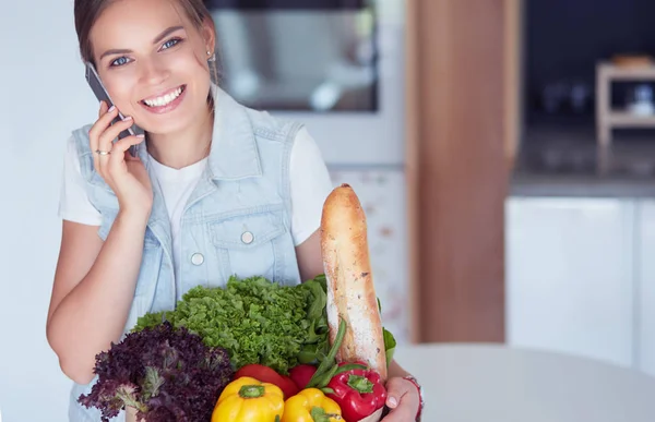 Smiling woman with mobile phone holding shopping bag in kitchen — Stock Photo, Image