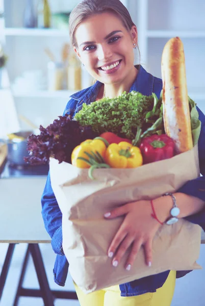 Sebzeli alışveriş poşeti taşıyan genç bir kadın. Mutfakta duruyor.