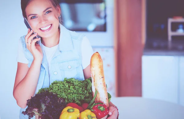 Smiling woman with mobile phone holding shopping bag in kitchen — Stock Photo, Image