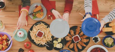 Ahşap masada otururken birlikte yemek yiyen bir grup insanın üst düzey görüntüsü. Masada yemek var. İnsanlar fast food yiyor..