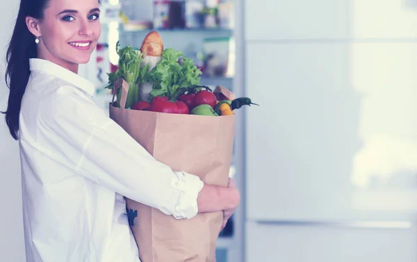 Mujer joven sosteniendo bolsa de la compra de comestibles con verduras.De pie en la cocina. Mujer en la cocina mirando a la cámara — Foto de Stock