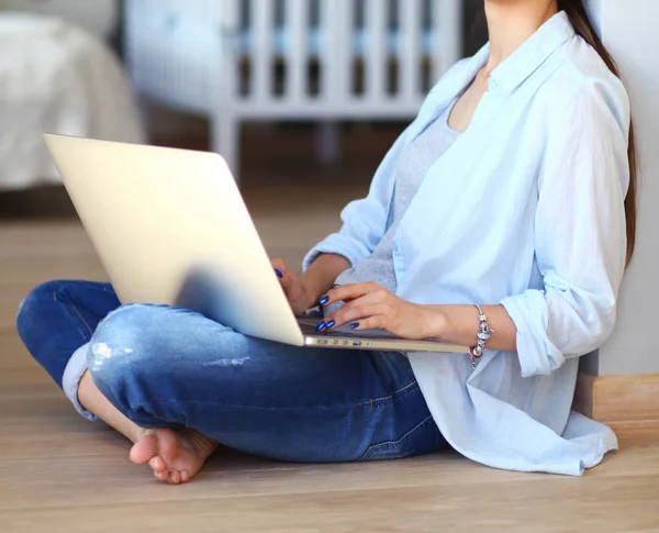 Ung kvinna som sitter på golvet nära barnens babysäng med laptop. Ung mamma — Stockfoto
