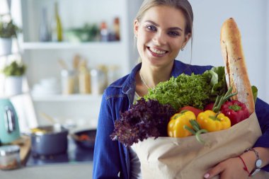 Sebzeli alışveriş poşeti taşıyan genç bir kadın. Mutfakta duruyor.