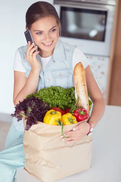 Улыбающаяся женщина с мобильным телефоном держит сумку для покупок на кухне — стоковое фото