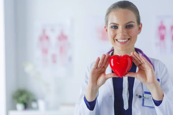 Young woman doctor holding a red heart, standing on hospital background — Stock Photo, Image