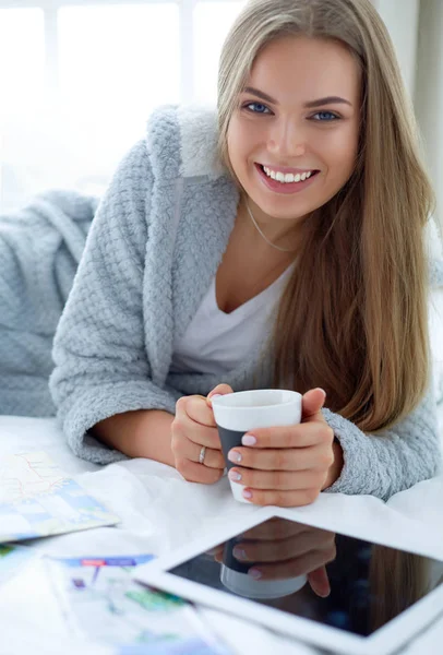 Avslappnad ung kvinna sitter på sängen med en kopp kaffe och digital tablett — Stockfoto