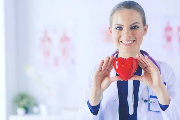 Young woman doctor holding a red heart, standing on hospital background — Stock Photo, Image