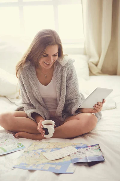 Uvolněná mladá žena sedící na posteli s šálkem kávy a digitální tablet — Stock fotografie