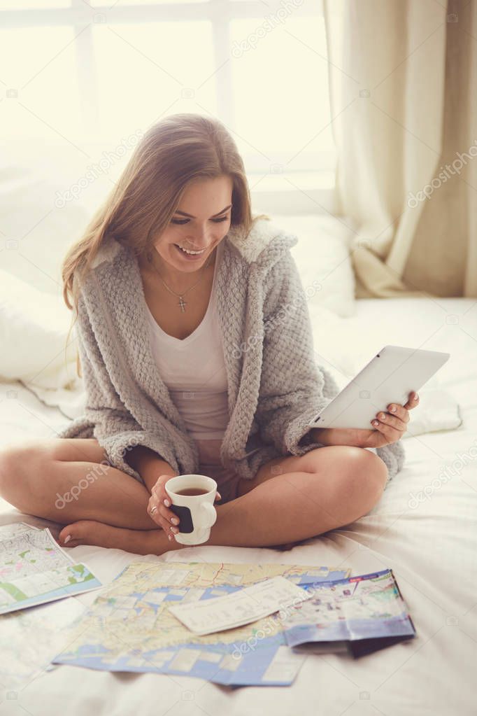 Relaxed young woman sitting on bed with a cup of coffee and digital tablet