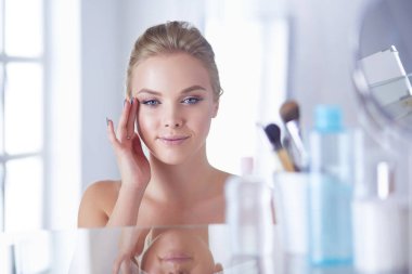 Bornozlu genç kadın banyo aynasına bakıyor.