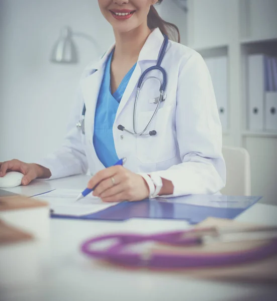 Mooie jonge glimlachende vrouwelijke arts zitten aan het bureau en schrijven. — Stockfoto