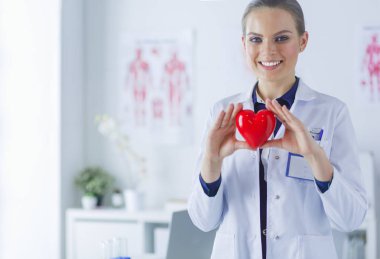 Kırmızı bir kalp taşıyan genç bir kadın doktor, hastane arka planında duruyor.