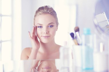 Bornozlu genç kadın banyo aynasına bakıyor.