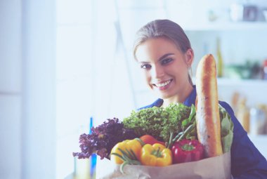 Sebzeli alışveriş poşeti taşıyan genç bir kadın. Mutfakta duruyor.