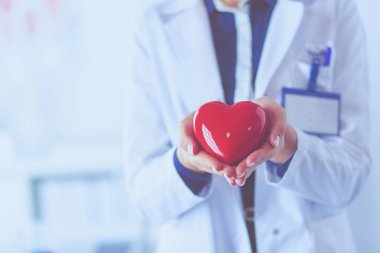 Kırmızı bir kalp taşıyan genç bir kadın doktor, hastane arka planında duruyor.