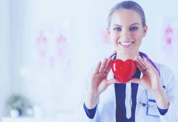 Young woman doctor holding a red heart, standing on hospital background — Stock Photo, Image