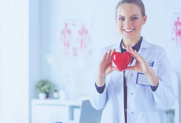 Mujer joven doctora sosteniendo un corazón rojo, de pie sobre el fondo del hospital — Foto de Stock