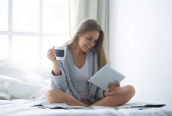 Avslappnad ung kvinna sitter på sängen med en kopp kaffe och digital tablett — Stockfoto