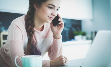 Genç kadın bir dizüstü bilgisayar kullanarak ve cep telefonuyla konuşurken mutfak masasında oturur.