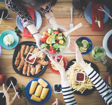Ahşap masada otururken birlikte yemek yiyen bir grup insanın üst düzey görüntüsü. Masada yemek var. İnsanlar fast food yiyor..