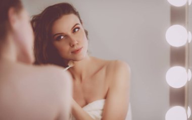 Genç bir kadın aynaya bakıyor ve makyaj yapıyor..