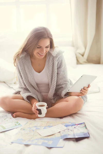 Avslappnad ung kvinna sitter på sängen med en kopp kaffe och digital tablett — Stockfoto