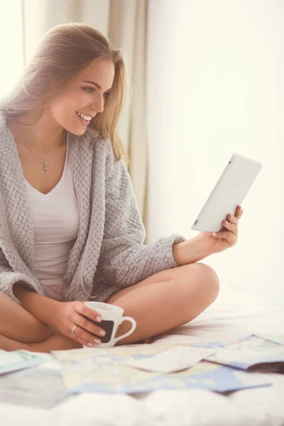 Relaxado jovem mulher sentada na cama com uma xícara de café e tablet digital — Fotografia de Stock