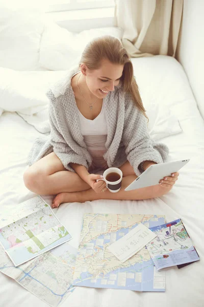 Mujer joven y relajada sentada en la cama con una taza de café y una tableta digital —  Fotos de Stock