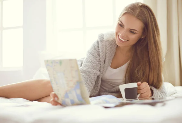 Relaxado jovem mulher sentada na cama com uma xícara de café e tablet digital — Fotografia de Stock