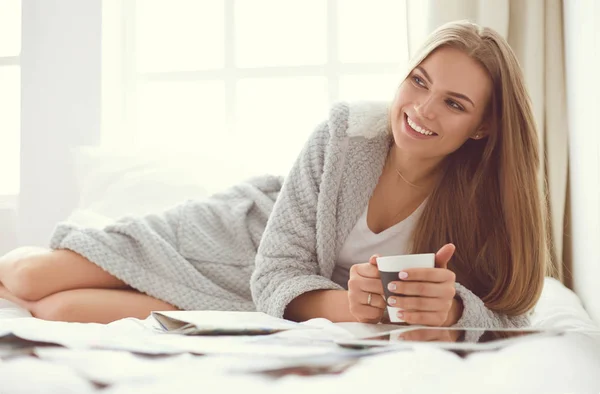 Relaxed young woman sitting on bed with a cup of coffee and digital tablet — Stock Photo, Image