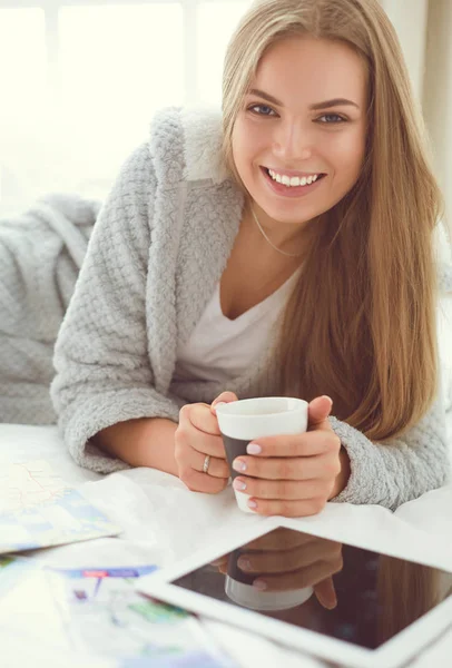 Розслаблена молода жінка сидить на ліжку з чашкою кави та цифровим планшетом — стокове фото